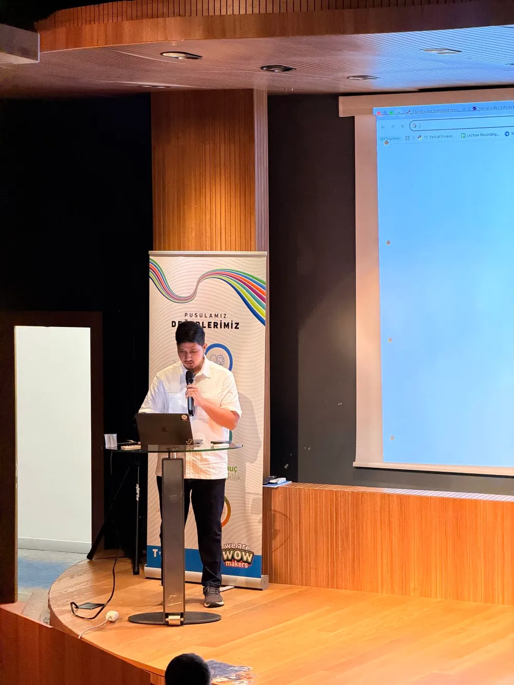Ali Mustufa Shaikh presenting Mirror with a microphone and laptop on a wood-panelled stage in Istanbul.