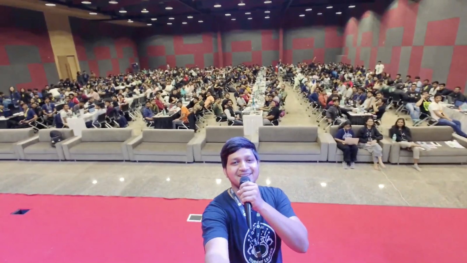 Ali Mustufa Shaikh on stage with a microphone, facing a packed hall of several hundred developers seated at long tables.